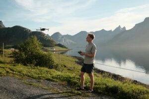 Mies seisoo ruohoisella järvenrantatien varrella ja ohjaa lennokkia kaukosäätimellä. Taustalla näkyy vuoria ja vettä kirkkaalla taivaalla.