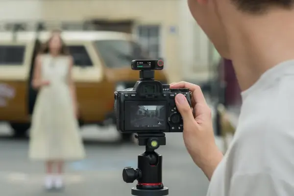 Henkilö käyttää DJI Mic 2 Dual Kit -laitteistoa kuvatessaan valkoiseen mekkoon pukeutunutta naista ulkona vintage-ajoneuvon vieressä; kameran näytöllä nainen näkyy tarkennettuna, vaikka hän näyttääkin epäselvältä taustalla.