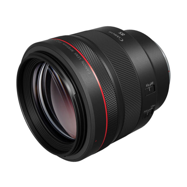 Canon RF 85mm f/1.2 L USM -objektiivi, jossa on musta runko, punainen rengasaksentti ja näkyvä tarkennusrengas, on kuvattu kuvakulmassa valkoista taustaa vasten.