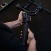 Henkilö kokoaa tai säätää DJI RS 4 Pro -käsikameragimbaalia mustalla pinnalla pitäen sen kahvasta kiinni molemmilla käsillä.